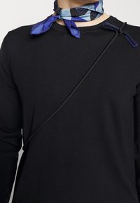 Haut noir côtelé à manches longues avec une sangle noire diagonale et des éléments métalliques avec accents bleus. La personne porte une écharpe à motifs bleus et noirs.