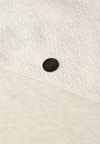 Světlá béžová látka s texturovaným geometrickým vzorem, obsahující černou kruhovou nášivku s logem a hladkou kontrastní oblast pod ní.