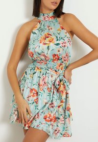 Žena oblečená v bezrukávových květinových šatech s halter krkem a uvázaným páskem v pase, na světle modrém pozadí.