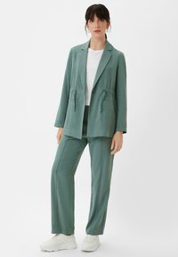 Mint green suit jacket and matching trousers, featuring a relaxed fit, adjustable drawstring waist, and sleek texture. Paired with white sneakers.