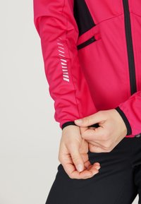 Lyserød atletisk jakke med sorte detaljer, med reflekterende striber på ærmet og en tætsiddende manchet. Lynlåslommer er synlige.