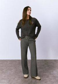 Suéter de canalé gris oscuro con un logo, combinado con pantalones acampanados gris claro y zapatillas crema. Texturas suaves, corte relajado y diseños distintos.