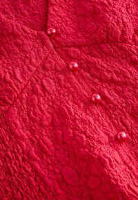 Material texturat roșu cu model de dantelă, având patru nasturi roșii lucioși ca accente, creând un efect vizual stratificat.