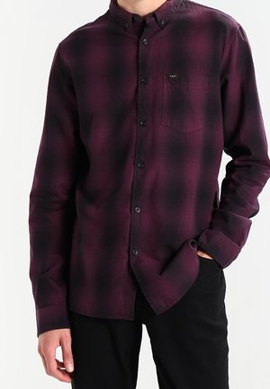 Homme portant une chemise à manches longues à carreaux violet et noir avec une poche frontale et un pantalon noir, une main dans la poche.