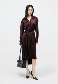 Bordowa sukienka z satyny z długimi rękawami, paskiem w talii i asymetrycznym dołem. Do tego czarne szpilki w szpic i granatowa, sztywna torebka.