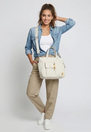 Jeune femme souriante, portant une chemise en denim, un débardeur blanc, un pantalon beige, des baskets blanches, tenant un grand sac cabas écru avec bandoulière.