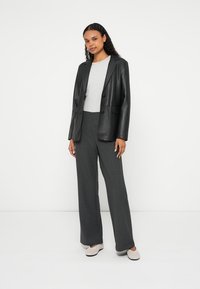 Γυναίκα όρθια, φοράει μαύρο δερμάτινο σακάκι, ανοιχτό γκρι τοπ, σκούρα γκρι παντελόνια με φαρδιά μπατζάκια και ανοιχτό μπεζ παπούτσια χωρίς κορδόνια, μπροστά σε λευκό φόντο.