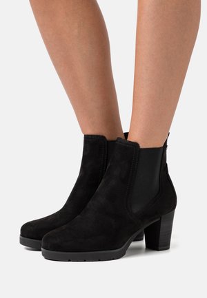 Jambes portant des bottines en daim noir avec des panneaux latéraux en élastique et des talons blocs empilés sur un fond clair.