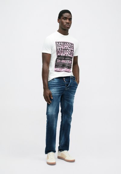 Weißes T-Shirt mit lila grafischem Text, kombiniert mit blauen Jeans aus Denim mit einem ausgewaschenen Design und beigen Sneakers mit braunen Akzenten.