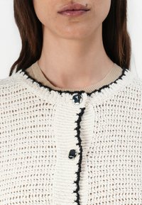 Cardigan in maglia di crema chiara con accenti neri e scollo rotondo, chiuso con un bottone lucido sul davanti.