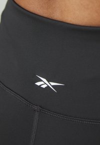 Sorte høyhåndte tights laget av elastisk stoff, med flatlock-sømmer og en liten hvit logo på midjebåndet.