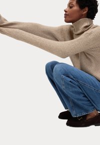 Beige geribbelde coltrui met lange mouwen, gecombineerd met blauwe wijde jeans. Het model hurkt en toont de ontspannen pasvorm en texturen.