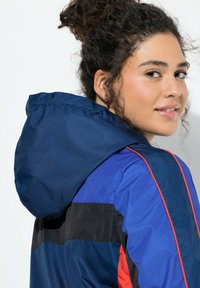 Modrá větrovka s kapucí, s červenými a černými akcenty na rukávech a texturovaným materiálem. Detail na záda a rameno.