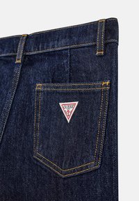 Dunkelblaue Denim-Jeans mit kontrastierenden gelben Nähten und einem dreieckigen Logo-Patch in Rot, Weiß und Blau auf der Gesäßtasche.