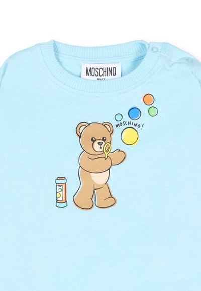 Jasnoniebieska koszulka dziecięca z misiem dmuchającym kolorowe bańki oraz napisem "MOSCHINO!" obok baniek.