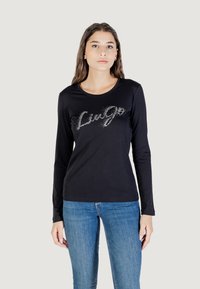 Una giovane donna indossa una maglietta nera a maniche lunghe con "Liu Jo" scritto in lettere scintillanti, abbinata a jeans blu, su uno sfondo neutro.