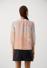 Femme aux cheveux tressés portant des boucles d'oreilles créoles, un blouse à patchwork pastel avec des manches 3/4, et une mini jupe en denim noir avec un ourlet effiloché, de dos.