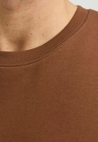Camisa de algodón marrón con cuello redondo, que presenta un escote acanalado y una textura suave, con costuras visibles a lo largo del cuello.