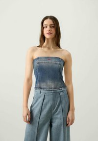 Jeans brez naramnic v svetlo modri barvi s prilegajočim dizajnom, ki ima majhen rdeč logotip na predelu prsi, v kombinaciji z hlačami širokega kroja visokega pasu.