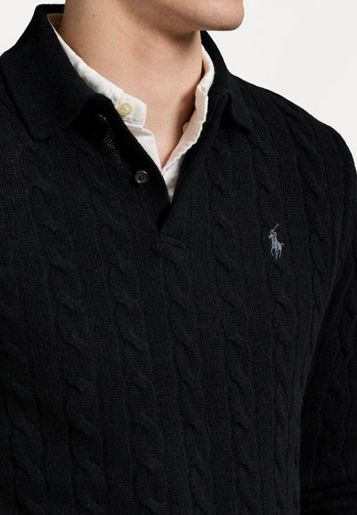 Pull en tricot noir à col, avec un logo sur la poitrine et superposé à une chemise blanche à boutons en dessous.