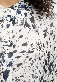 Άσπρο πουκάμισο με μοτίβο πιτσιλιών σε μπλε και μαύρο, διαθέτει μυτερό γιακά και κούμπωμα με κουμπιά, κατασκευασμένο από ελαφρύ ύφασμα με υφή.