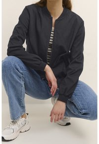 Giacca bomber leggera nera con colletto a coste, chiusura con zip e maniche lunghe, indossata sopra una maglietta a righe e jeans in denim blu.