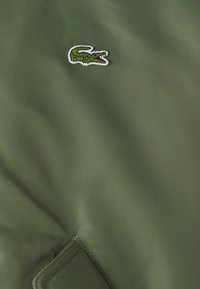 Material de exterior verde cu o textură netedă, având un mic logo brodat al unui crocodil în alb și roșu. Detaliu cu patch vizibil.