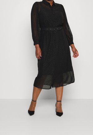 Kvinde iført en sort midikjole med gennemsigtige lange ærmer, et bælte om taljen og sorte hæle med remme om anklerne mod en ensfarvet baggrund.