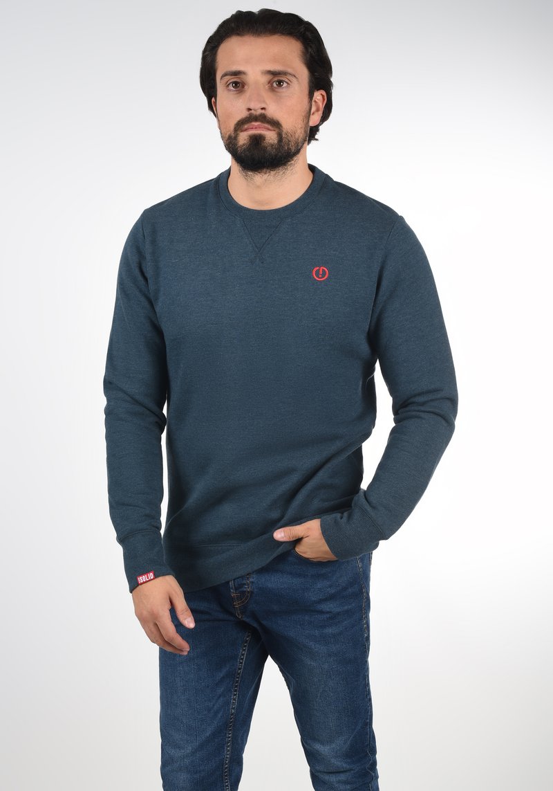 Donkerblauwe sweatshirt van zachte stof, met een ronde hals, lange mouwen en een klein rood logo op de borst. Draagt blauw spijkerbroek.