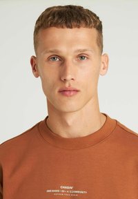 Hnědá mikina s kulatým výstřihem, žebrovaným límcem a bílým potiskem. Model má krátké, světle hnědé vlasy a modré oči.