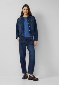 Jeansjack met knoopsluiting aan de voorkant en twee borstzakken, gecombineerd met een blauwe ronde hals t-shirt en donkerblauwe loszittende jeans. Bruine loafers.