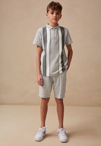 Vaaleanharmaa poolopaita, jossa on pystysuoria tummanharmaita ja valkoisia raitoja, yhdistetty samanlaisiin shortseihin. Kangas näyttää pehmeältä, rennon tyylikäs asu, valkoiset lenkkarit jalassa.