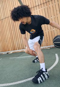 Camiseta negra con gráfico de baloncesto, pantalones cortos blancos con detalles en negro y zapatillas negras con grandes letras "AIR" en los lados.