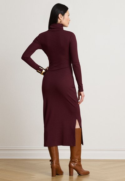 Femme portant une longue robe col roulé bordeaux avec une fente latérale, des bottes marron hautes à talons et un bracelet doré, debout contre un mur uni.
