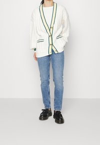 Vit kabelstickad cardigan med grön och gul randig detalj, med gula knappar och fickor, matchad med blå jeans och svarta skor.