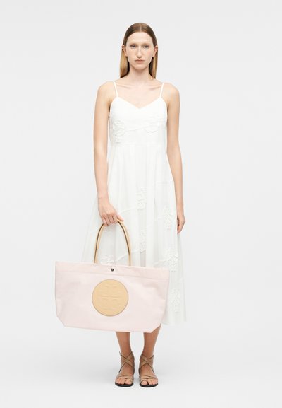 Γυναίκα με λευκό αμάνικο φόρεμα που κρατά μεγάλη τσάντα ώμου απαλού ροζ χρώματος με καφέ κυκλικό λογότυπο, φοράει σανδάλια με λουράκια, στέκεται μπροστά σε λευκό φόντο.