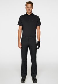 Svart pikétröja med korta ärmar, i kombination med svarta byxor. Modellen bär en svart handske och svarta skor med vita accenter.