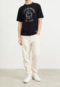 Persona che indossa una t-shirt nera con grafica del Super Bowl e pantaloni beige, in piedi su un pavimento chiaro contro uno sfondo minimale.
