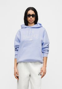 Φαρδιά μωβ μπλούζα με κουκούλα και μπροστινή τσέπη με τη φράση "NEW BALANCE ATHLETICS". Συνδυάζεται με γκρι φόρμα με λογότυπο.