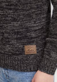 Stickad tröja i mörkgrått och svart med ribbat mönster. Har en brun läderlapp med "Solid" och dekorativa detaljer.