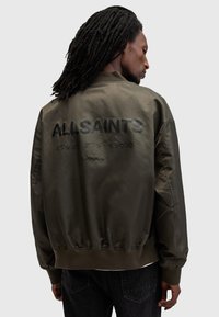 Olivenfarvet bomberjakke, mat finish, ribbede manchetter og kant, med stor sort tekst "ALLSAINTS" på ryggen og yderligere grafiske detaljer.