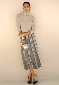 Frau in beigem Rollkragenpullover, schwarz-weiß gemustertem Rock, schwarzen High Heels, hält eine geometrische goldene Clutch, steht vor einer beigen Wand.