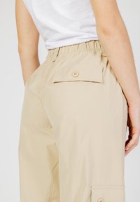 Pantaloni cargo beige con vita elastica, dotati di una tasca posteriore con chiusura a bottone e tasche cargo laterali. Tessuto dalla superficie liscia.