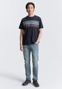 Marineblaues T-Shirt mit kurzen Ärmeln und horizontalen Streifen in Hellblau und Rot. Getragen mit hellblauen Jeans und dunklen Schuhen.