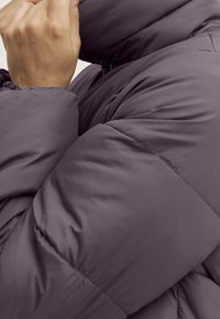 Osoba poprawiająca kołnierz fioletowej, pikowanej zimowej kurtki z elastycznymi mankietami, pokazana w zbliżeniu.