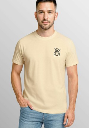 Beige Baumwoll-T-Shirt mit einer schwarzen Linzeichnung eines Teddybären auf der linken Brust. Kurzärmlig und mit Rundhalsausschnitt.
