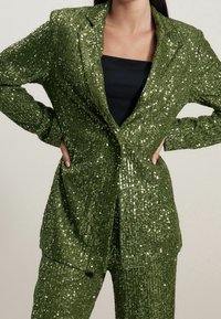 Groene sequin blazer met een gestructureerde afwerking, voorzien van een enkele knoopsluiting en twee voorzakken, gecombineerd met een zwarte top eronder.