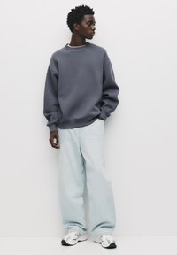 Grå sweatshirt med rund hals, oversized fit, parret med lyseblå brede bukser og sølvfarvede sneakers. Glat stof, afslappet silhuet.