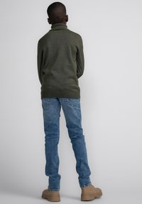 Maglione a collo alto in verde oliva a maglia, jeans in denim blu e calzature beige. Il maglione ha un design aderente, mentre i jeans sono a taglio slim.