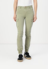 Femme portant un pantalon cargo jogger vert clair avec des chevilles resserrées et des baskets noires avec des rayures blanches, se tenant devant un fond blanc.